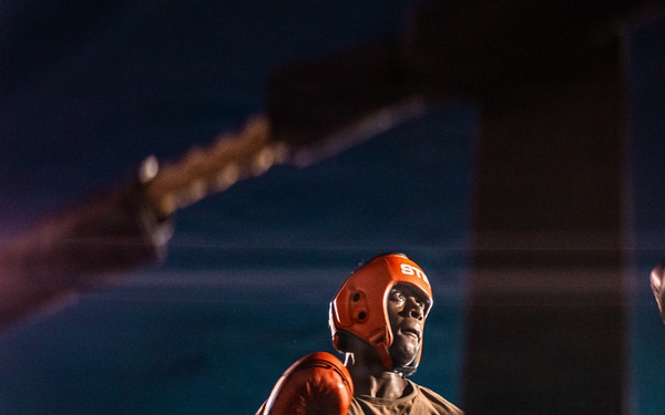 Two boxers are engaged in a match inside a boxing ring under bright artificial lights at night. The central fighter wears an orange/red protective headgear and orange boxing gloves, facing an opponent in a dark shirt with 'Quartermaster' text visible on the back. The dramatic low-angle shot and ring ropes frame the intense athletic confrontation.