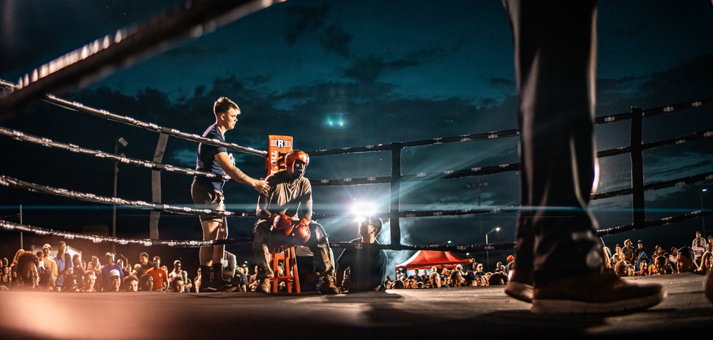 An outdoor boxing event taking place at night, with a boxer seated on a stool in their corner between rounds while a trainer or cornerman attends to them. The ring is illuminated by bright spotlights against a dramatic dark blue cloudy sky. A large crowd of spectators surrounds the ring, suggesting a well-attended public event.