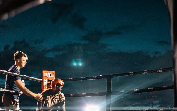 An outdoor boxing event taking place at night, with a boxer seated on a stool in their corner between rounds while a trainer or cornerman attends to them. The ring is illuminated by bright spotlights against a dramatic dark blue cloudy sky. A large crowd of spectators surrounds the ring, suggesting a well-attended public event.