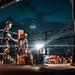 An outdoor boxing event taking place at night, with a boxer seated on a stool in their corner between rounds while a trainer or cornerman attends to them. The ring is illuminated by bright spotlights against a dramatic dark blue cloudy sky. A large crowd of spectators surrounds the ring, suggesting a well-attended public event.