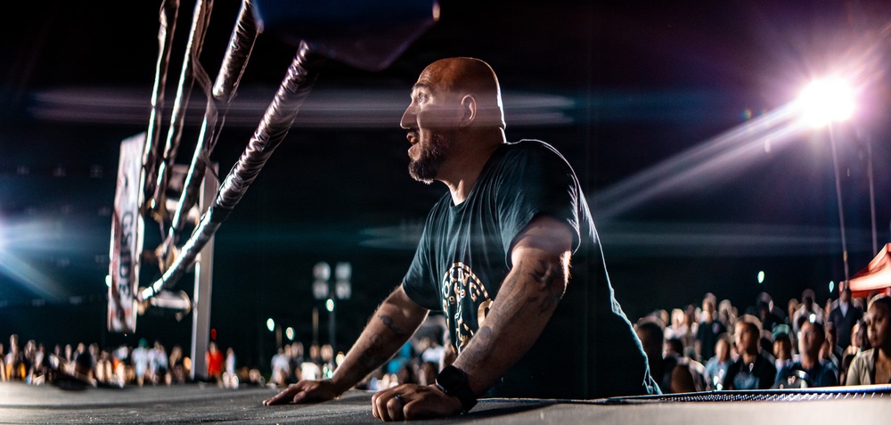 A bald man with a beard and tattoos leans against the edge of a professional wrestling ring, appearing to shout or react intensely. The scene is set at a nighttime outdoor or indoor arena event with dramatic stage lighting and a large crowd visible in the background. The low-angle shot emphasizes the ring ropes and corner post in the foreground.