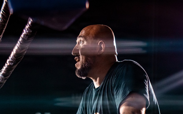 A bald man with a beard and tattoos leans against the edge of a professional wrestling ring, appearing to shout or react intensely. The scene is set at a nighttime outdoor or indoor arena event with dramatic stage lighting and a large crowd visible in the background. The low-angle shot emphasizes the ring ropes and corner post in the foreground.