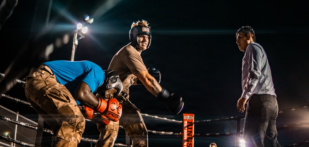 A nighttime boxing or combat sports match taking place in a ring under bright stadium lights. Two fighters are engaged in a clinch or grappling exchange, with one wearing a headguard and boxing gloves, while a third person — likely a referee or official — stands nearby observing. The dramatic low-angle lighting and dark background create a high-contrast, atmospheric scene.