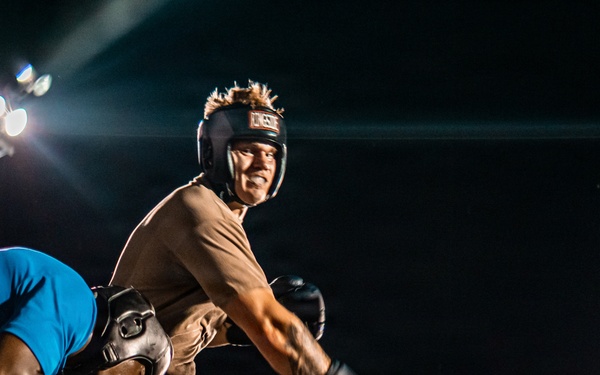 A nighttime boxing or combat sports match taking place in a ring under bright stadium lights. Two fighters are engaged in a clinch or grappling exchange, with one wearing a headguard and boxing gloves, while a third person — likely a referee or official — stands nearby observing. The dramatic low-angle lighting and dark background create a high-contrast, atmospheric scene.
