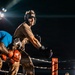 A nighttime boxing or combat sports match taking place in a ring under bright stadium lights. Two fighters are engaged in a clinch or grappling exchange, with one wearing a headguard and boxing gloves, while a third person — likely a referee or official — stands nearby observing. The dramatic low-angle lighting and dark background create a high-contrast, atmospheric scene.