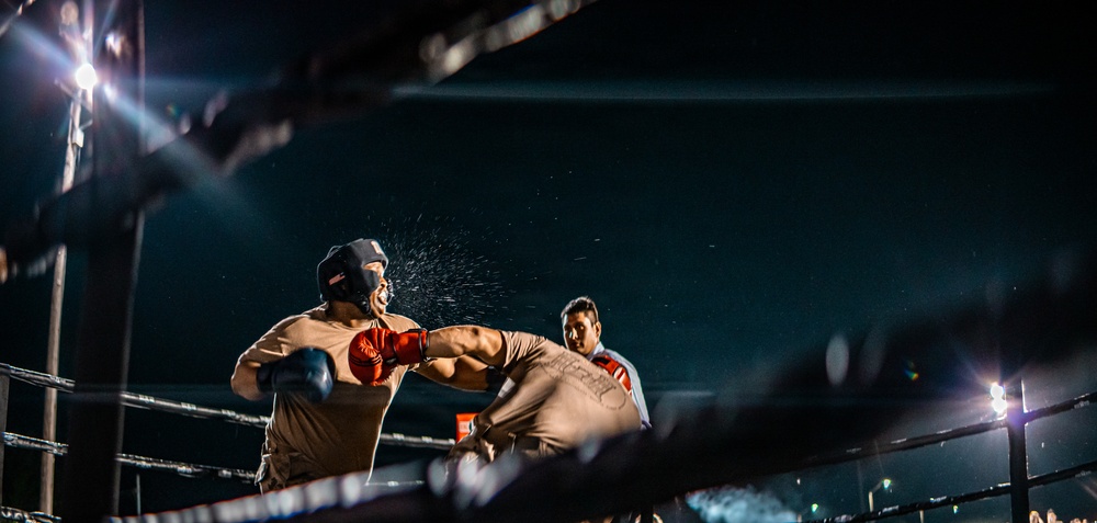 A boxing match is captured mid-action in an outdoor or open-air ring at night, with one fighter landing a punch on the other, causing visible sweat or water droplets to spray from the impact. A referee is visible in the background monitoring the bout. The dramatic low-angle shot and ring-side lighting create a high-contrast, intense atmosphere.