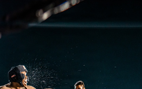 A boxing match is captured mid-action in an outdoor or open-air ring at night, with one fighter landing a punch on the other, causing visible sweat or water droplets to spray from the impact. A referee is visible in the background monitoring the bout. The dramatic low-angle shot and ring-side lighting create a high-contrast, intense atmosphere.