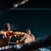 A boxing match is captured mid-action in an outdoor or open-air ring at night, with one fighter landing a punch on the other, causing visible sweat or water droplets to spray from the impact. A referee is visible in the background monitoring the bout. The dramatic low-angle shot and ring-side lighting create a high-contrast, intense atmosphere.