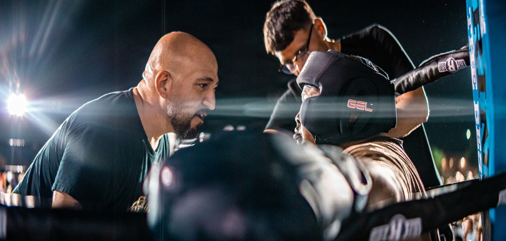 The image depicts an intense corner scene in a boxing match, with a bald trainer or cornerman leaning in close to speak with a boxer between rounds. A second person, likely another cornerman, is visible in the background attending to the boxer's gloves or wraps. The scene is dramatically lit with bright ring lights against a dark arena background.