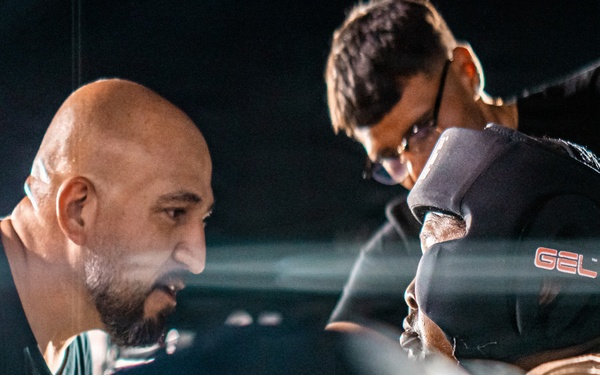 The image depicts an intense corner scene in a boxing match, with a bald trainer or cornerman leaning in close to speak with a boxer between rounds. A second person, likely another cornerman, is visible in the background attending to the boxer's gloves or wraps. The scene is dramatically lit with bright ring lights against a dark arena background.