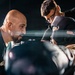 The image depicts an intense corner scene in a boxing match, with a bald trainer or cornerman leaning in close to speak with a boxer between rounds. A second person, likely another cornerman, is visible in the background attending to the boxer's gloves or wraps. The scene is dramatically lit with bright ring lights against a dark arena background.