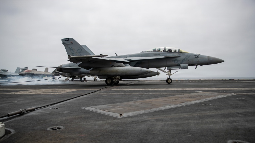 A U.S. Navy carrier-based jet aircraft, marked with the number 107, is landing or rolling out on what appears to be an aircraft carrier flight deck. The arresting wire is visible on the deck, consistent with a carrier arrested landing. Additional aircraft and smoke from jet exhaust are visible in the background.