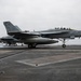 A U.S. Navy carrier-based jet aircraft, marked with the number 107, is landing or rolling out on what appears to be an aircraft carrier flight deck. The arresting wire is visible on the deck, consistent with a carrier arrested landing. Additional aircraft and smoke from jet exhaust are visible in the background.