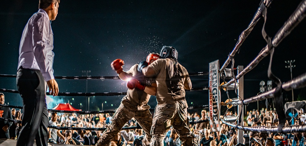An outdoor nighttime boxing match between two fighters wearing military camouflage uniforms and protective headgear, with red boxing gloves. A referee in formal attire (white dress shirt and black trousers) oversees the bout from the left side of the ring. The event is well-attended, with a large crowd of spectators visible in the background surrounding the illuminated ring.
