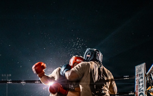 An outdoor nighttime boxing match between two fighters wearing military camouflage uniforms and protective headgear, with red boxing gloves. A referee in formal attire (white dress shirt and black trousers) oversees the bout from the left side of the ring. The event is well-attended, with a large crowd of spectators visible in the background surrounding the illuminated ring.