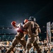An outdoor nighttime boxing match between two fighters wearing military camouflage uniforms and protective headgear, with red boxing gloves. A referee in formal attire (white dress shirt and black trousers) oversees the bout from the left side of the ring. The event is well-attended, with a large crowd of spectators visible in the background surrounding the illuminated ring.