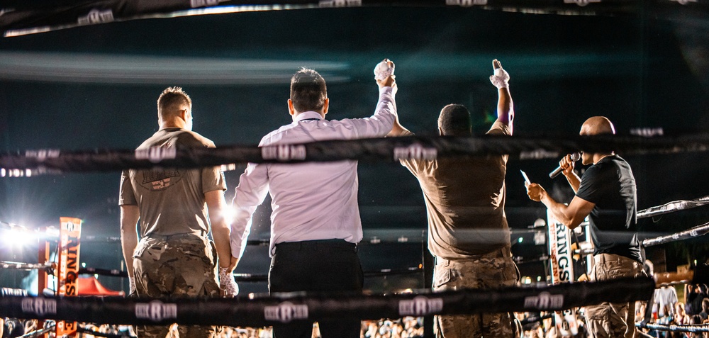 A boxing match conclusion is captured from behind the ring ropes, showing a referee raising the arm of the winner in a victory declaration. Two fighters in military camouflage uniforms stand in the ring alongside the referee in a white shirt and black pants. An announcer holding a microphone stands to the right, addressing the crowd visible in the background.