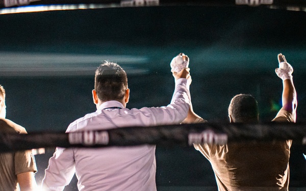 A boxing match conclusion is captured from behind the ring ropes, showing a referee raising the arm of the winner in a victory declaration. Two fighters in military camouflage uniforms stand in the ring alongside the referee in a white shirt and black pants. An announcer holding a microphone stands to the right, addressing the crowd visible in the background.