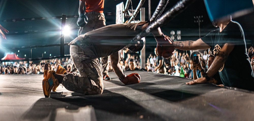 A performer in camouflage pants and orange sneakers is kneeling and bowing down on a stage in front of a large crowd at what appears to be an outdoor concert or event. The low-angle shot captures the stage edge with audience members reaching toward the performer. Stage lighting and rigging are visible in the background.