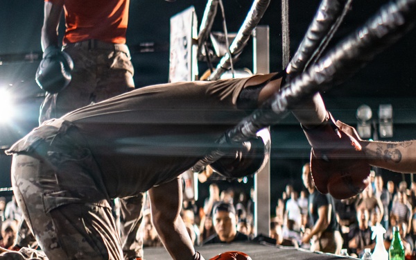 A performer in camouflage pants and orange sneakers is kneeling and bowing down on a stage in front of a large crowd at what appears to be an outdoor concert or event. The low-angle shot captures the stage edge with audience members reaching toward the performer. Stage lighting and rigging are visible in the background.