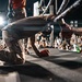 A performer in camouflage pants and orange sneakers is kneeling and bowing down on a stage in front of a large crowd at what appears to be an outdoor concert or event. The low-angle shot captures the stage edge with audience members reaching toward the performer. Stage lighting and rigging are visible in the background.
