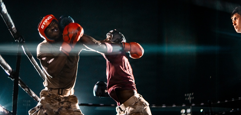 Two boxers are engaged in an active match inside a boxing ring, with one fighter delivering a punch to the other. Both fighters are wearing headgear and boxing gloves, and appear to be dressed in military-style camouflage pants, suggesting this may be a military boxing event. The image is captured mid-action under dramatic spotlight lighting against a dark background.