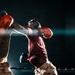 Two boxers are engaged in an active match inside a boxing ring, with one fighter delivering a punch to the other. Both fighters are wearing headgear and boxing gloves, and appear to be dressed in military-style camouflage pants, suggesting this may be a military boxing event. The image is captured mid-action under dramatic spotlight lighting against a dark background.
