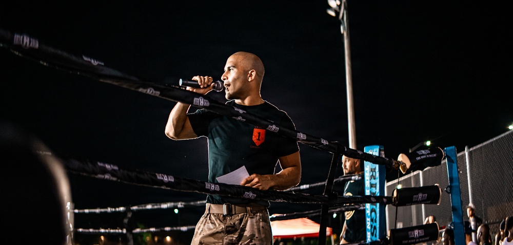 A bald man wearing a black t-shirt with a red logo and camouflage pants stands inside or beside a boxing/MMA ring at night, speaking into a microphone. He appears to be an announcer or emcee at an outdoor combat sports event. The scene is illuminated by stadium lighting against a dark night sky.