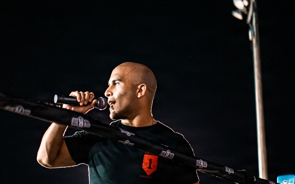 A bald man wearing a black t-shirt with a red logo and camouflage pants stands inside or beside a boxing/MMA ring at night, speaking into a microphone. He appears to be an announcer or emcee at an outdoor combat sports event. The scene is illuminated by stadium lighting against a dark night sky.