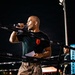 A bald man wearing a black t-shirt with a red logo and camouflage pants stands inside or beside a boxing/MMA ring at night, speaking into a microphone. He appears to be an announcer or emcee at an outdoor combat sports event. The scene is illuminated by stadium lighting against a dark night sky.
