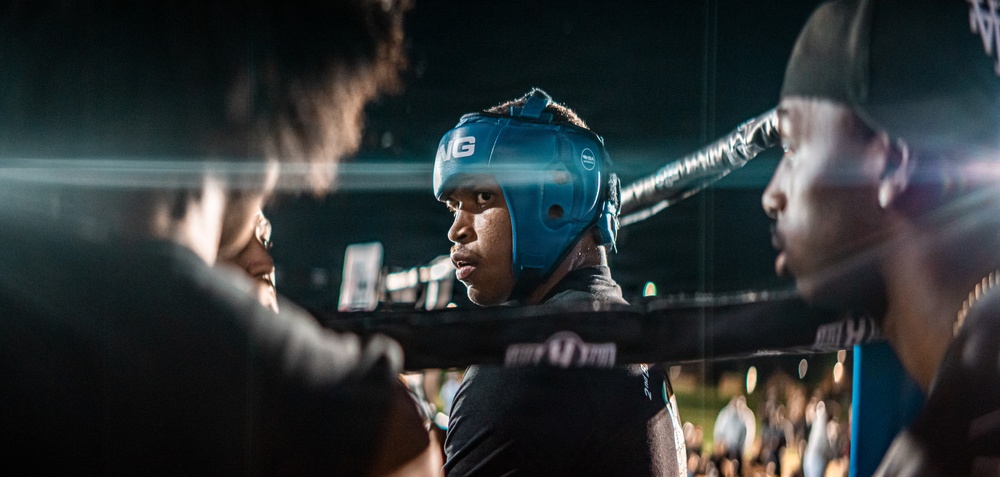 A boxer wearing a blue protective headgear is positioned at the corner of a boxing ring, surrounded by corner team members between rounds. The scene appears to be an active boxing event with an audience visible in the background. The dramatic lighting and close framing create an intense, intimate atmosphere.