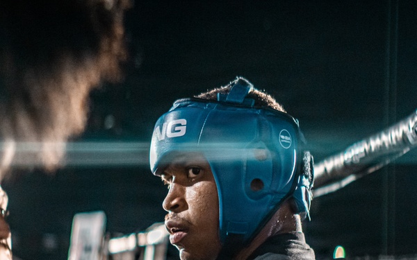 A boxer wearing a blue protective headgear is positioned at the corner of a boxing ring, surrounded by corner team members between rounds. The scene appears to be an active boxing event with an audience visible in the background. The dramatic lighting and close framing create an intense, intimate atmosphere.
