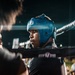 A boxer wearing a blue protective headgear is positioned at the corner of a boxing ring, surrounded by corner team members between rounds. The scene appears to be an active boxing event with an audience visible in the background. The dramatic lighting and close framing create an intense, intimate atmosphere.
