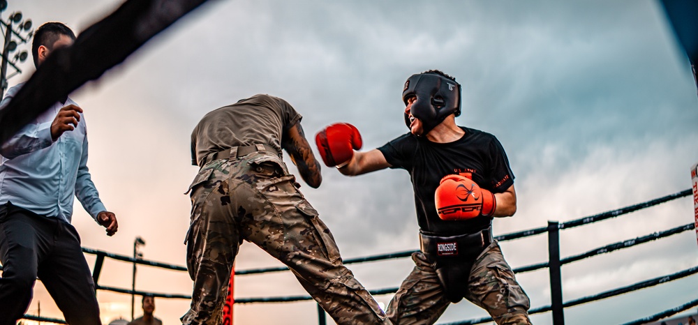 Two fighters wearing military camouflage pants are engaged in an outdoor boxing match inside a ring. One fighter in a black shirt with red-and-orange gloves and a black headguard is throwing or receiving a punch, while the other fighter faces away from the camera. A referee or official in a white shirt is visible on the left side of the ring.