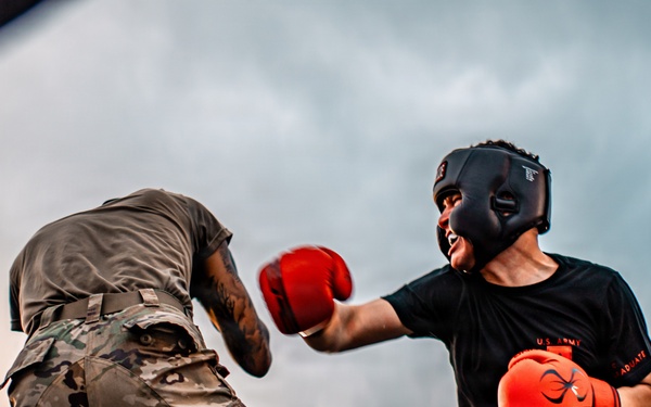 Two fighters wearing military camouflage pants are engaged in an outdoor boxing match inside a ring. One fighter in a black shirt with red-and-orange gloves and a black headguard is throwing or receiving a punch, while the other fighter faces away from the camera. A referee or official in a white shirt is visible on the left side of the ring.