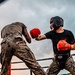 Two fighters wearing military camouflage pants are engaged in an outdoor boxing match inside a ring. One fighter in a black shirt with red-and-orange gloves and a black headguard is throwing or receiving a punch, while the other fighter faces away from the camera. A referee or official in a white shirt is visible on the left side of the ring.