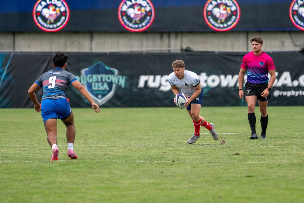 A rugby match is being played at a Rugbytown USA venue, likely in Glendale, Colorado. A player in a white jersey and red socks carries the ball while being approached by an opposing player wearing number 9 in a gray and blue uniform. A match official in a pink and purple gradient jersey observes the play from the right side.