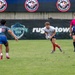 A rugby match is being played at a Rugbytown USA venue, likely in Glendale, Colorado. A player in a white jersey and red socks carries the ball while being approached by an opposing player wearing number 9 in a gray and blue uniform. A match official in a pink and purple gradient jersey observes the play from the right side.