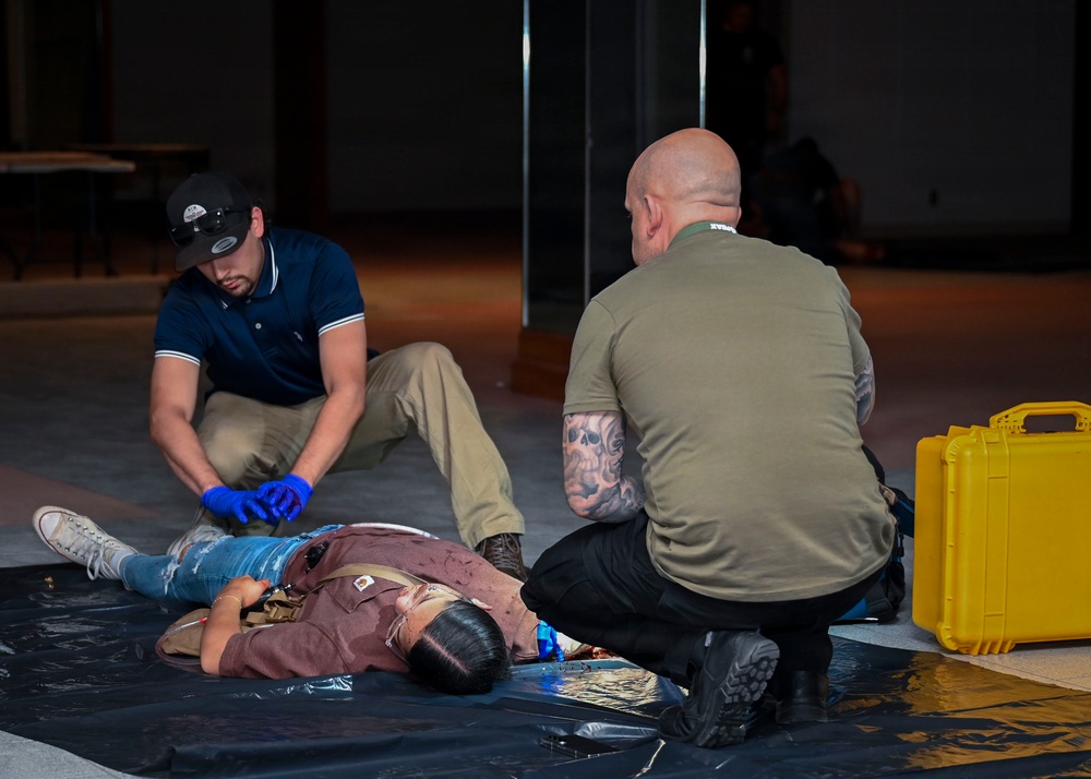 Two individuals are attending to a person lying on a black tarp on the floor, appearing to be a medical training or emergency response drill scenario. One person wearing blue latex gloves and a navy polo shirt is positioned near the subject's lower body, while a bald individual with visible tattoos in an olive shirt kneels near the subject's upper body. A yellow hard-shell equipment case is visible to the right, consistent with a first aid or tactical medical training exercise.