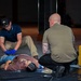 Two individuals are attending to a person lying on a black tarp on the floor, appearing to be a medical training or emergency response drill scenario. One person wearing blue latex gloves and a navy polo shirt is positioned near the subject's lower body, while a bald individual with visible tattoos in an olive shirt kneels near the subject's upper body. A yellow hard-shell equipment case is visible to the right, consistent with a first aid or tactical medical training exercise.