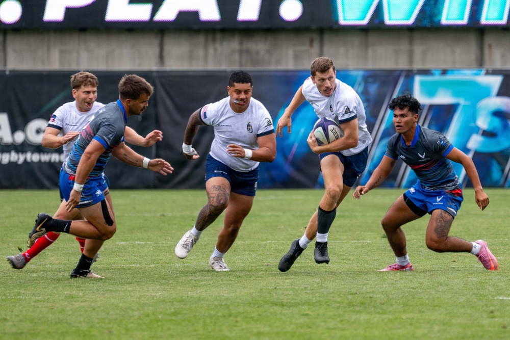 A rugby match is in progress on a grass field, with one player in a white jersey carrying the ball while being pursued by teammates and defended by opponents in gray and blue uniforms. The action is dynamic, capturing a mid-play moment with multiple players in motion. Advertising banners and what appears to be a stadium scoreboard are visible in the background.
