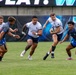 A rugby match is in progress on a grass field, with one player in a white jersey carrying the ball while being pursued by teammates and defended by opponents in gray and blue uniforms. The action is dynamic, capturing a mid-play moment with multiple players in motion. Advertising banners and what appears to be a stadium scoreboard are visible in the background.