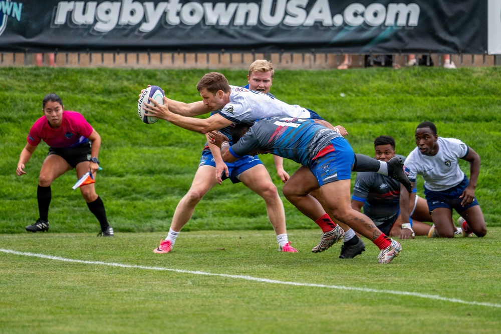 A rugby match is being played at a venue associated with rugbytownUSA.com, visible on the banner in the background. A ball carrier in a white jersey is being tackled by an opponent in a blue and red uniform near the try line. A female referee in a pink shirt and black shorts observes the play from the left sideline.