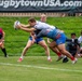 A rugby match is being played at a venue associated with rugbytownUSA.com, visible on the banner in the background. A ball carrier in a white jersey is being tackled by an opponent in a blue and red uniform near the try line. A female referee in a pink shirt and black shorts observes the play from the left sideline.