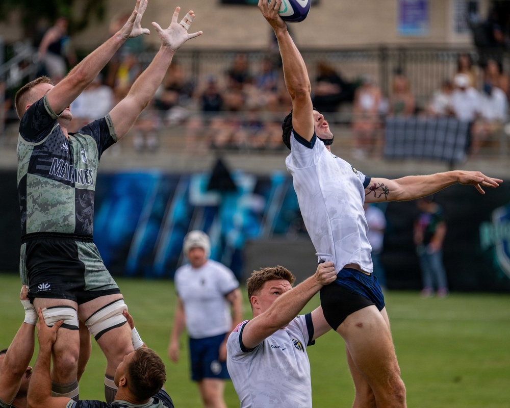 A rugby lineout contest is taking place between two teams, with players leaping to contest possession of the ball at height. One team is wearing a military-themed 'Marines' camouflage jersey in dark tones, while the opposing team wears white and navy uniforms. A crowd of spectators is visible in the stadium stands in the background.