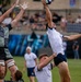 A rugby lineout contest is taking place between two teams, with players leaping to contest possession of the ball at height. One team is wearing a military-themed 'Marines' camouflage jersey in dark tones, while the opposing team wears white and navy uniforms. A crowd of spectators is visible in the stadium stands in the background.