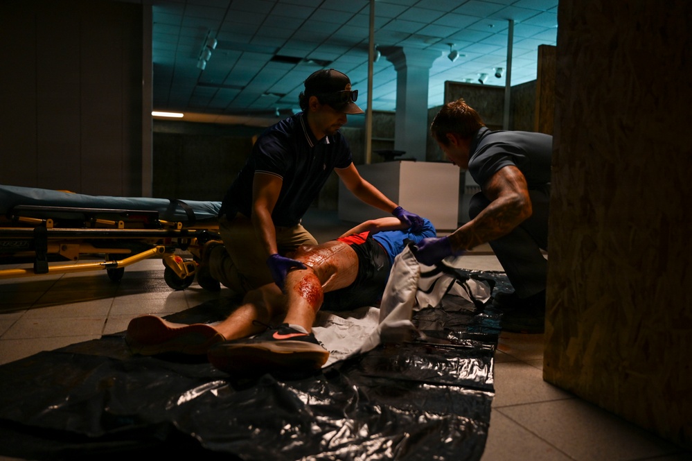 Two individuals wearing purple medical gloves are attending to a person lying on the floor in a dimly lit indoor space. The patient appears to have visible injuries with blood on their legs. A stretcher/gurney is visible in the background, suggesting a medical response scenario.