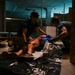 Two individuals wearing purple medical gloves are attending to a person lying on the floor in a dimly lit indoor space. The patient appears to have visible injuries with blood on their legs. A stretcher/gurney is visible in the background, suggesting a medical response scenario.