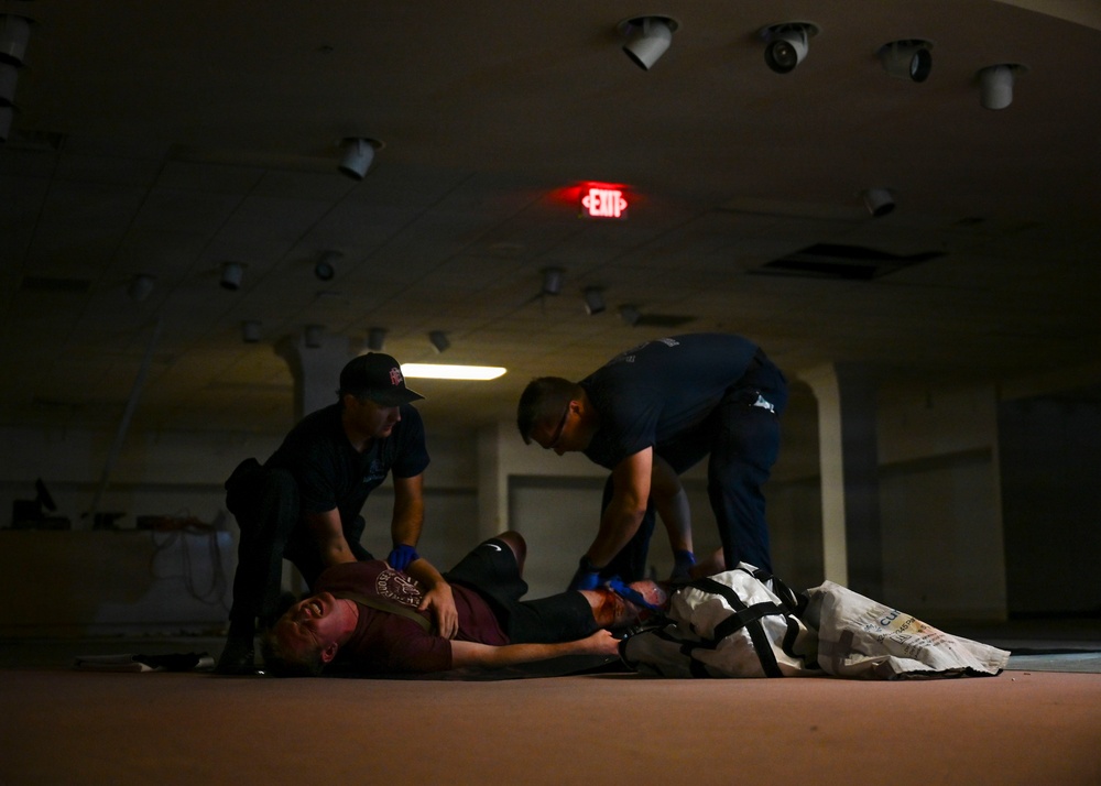 Two individuals wearing blue gloves and dark uniforms are attending to a person lying on the floor in a dimly lit indoor space. The scene appears to be a medical training exercise or emergency response drill, with what looks like simulated injuries and medical equipment visible. The setting appears to be a large, mostly empty interior space such as a warehouse or vacant commercial building.