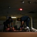 Two individuals wearing blue gloves and dark uniforms are attending to a person lying on the floor in a dimly lit indoor space. The scene appears to be a medical training exercise or emergency response drill, with what looks like simulated injuries and medical equipment visible. The setting appears to be a large, mostly empty interior space such as a warehouse or vacant commercial building.